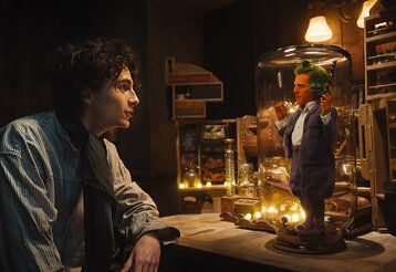 Ein junger Mann sitzt in einem dunklen Zimmer und spricht mit einem sehr kleinen Mann, der auf einem Tisch, unter einer Glasglocke steht