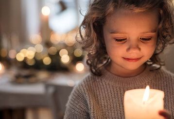 ein Mädchen hält eine Kerze vor sich und schaut sie an, im Hintergund weihnachtliche Lichter