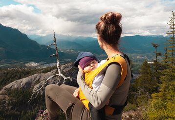 Frau mit Baby in Babytrage sitzt auf einem Berg und guckt in die Landschaft, das Baby guckt in die Kamera