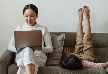 Frau sitzt mit Laptop auf dem Sofa, daneben sitzt ihr Kind verkehrt herum und streckt die Beine in die Höhe