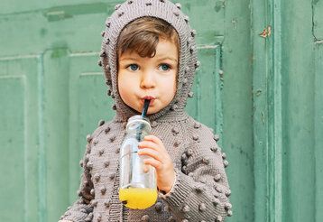 Kind mit Kapuzenpulli trinkt mit Strohhalm aus einer Trinkflasche, im Hintergrund eine grüne Tür