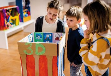 Drei Kinder stehen im Museum und gucken sich ein Kunstwerk aus Pappe von einem Kind an.
