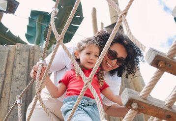 fröhliche Mutter ermutigt ihre Tochter, gemeinsam auf einer Strickleiter im öffentlichen Park zu spielen