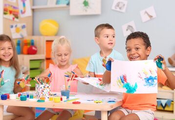 4 Kinder sitzen an einem Kindertisch, bemalenihre Hände und machen Handabdrücke auf Papier, ein Junge hält sein Bild in die Kamera