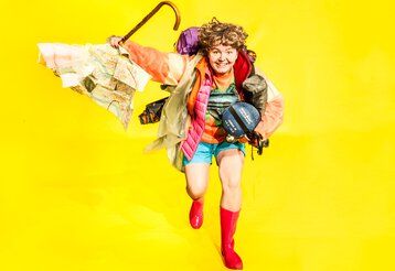 Eine Frau in Regenkleidung mit Wanderrucksack und Schirm rennt lachend Richtung Kamera.