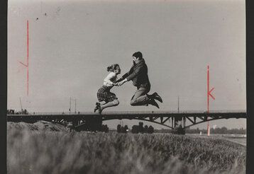 Ein schwarz-weiß Bild zeigt eine Frau und einen Mann die sich an den Händen haltend ansehen und auf einer Wiese vor einer Brücke in die Luft springen