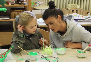 ein Mädchen und eine junge Frau sitzen im Atelier und bemalen ein Ton-Objekt