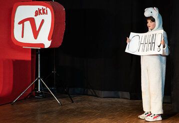 Ein Kind im Bärenkostüm steht auf einer Bühne für Aufnahmen von Akki TV.
