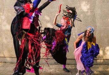 Drei Tänzerinnen und Tänzer bewegen sich vor einer Steinmauer, eingehüllt in verschiedene Stoffe
