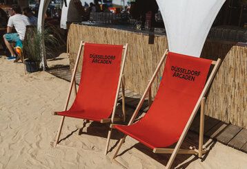 Zwei rote Liegestühle stehen im Sand eines Beachclubs.