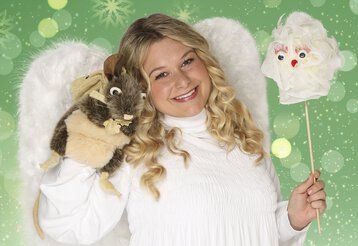 Ein blondes Mädchen lächelt als Engel verkleidet in die Kamera und hält eine Maus-Handpuppe in der Hand.