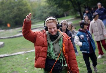 Eine Frau mit vielen Kabeln um den Hals und Kopfhörern auf den Ohren läuft lächelnd vor einer Gruppe Kinder über eine Wiese und hebt dabei einen Arm.