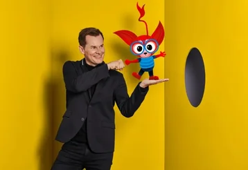 Malte Arkona steht vor einer gelben Wand mit der Kunstfigur Mezzo auf seiner Hand.