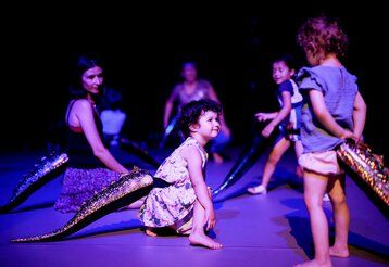 Kleine Kinder bewegen sich mit auf den Rücken geschnallten Reptilienschwänzen über die Bühne.