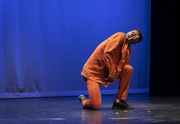 Ein Mann in orangefarbener Kleidung kniet mit geschlossenen Augen auf der Bühne.