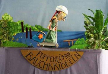 Bühnenkulisse der Puppenbühne Bauchkribbeln mit Kasperle auf einem Roller mit Helm und Sonnenbrille.