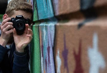 Ein junger Mann fotografiert Graffiti an einer Hauswand lehnend.