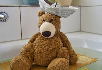 Ein Teddybär mit Papierhut sitzt auf einem Stück Holz in einer vollen Badewanne