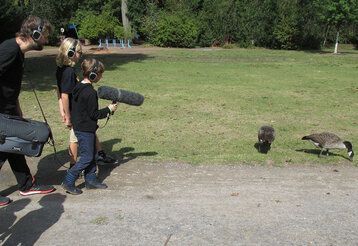 Zwei Kinder und ein Tontechniker nehmen mit Mikrofon und Headsets die Geräusche zweier Gänse in einem Park auf.