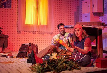 Eine Frau und ein Mann sitzen auf dem Boden der Bühne in der Kulisse einer Küche.