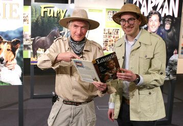 Zwei Männer stehen  in Safari-Anzügen im Filmmuseum vor alten Filmplakaten, in der Hand ein Buch über Lassie