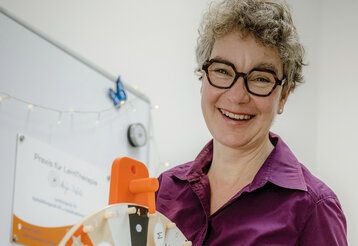 Frau mit auffälliger Brille lacht in die Kamera, im Hintergrund ein White Board