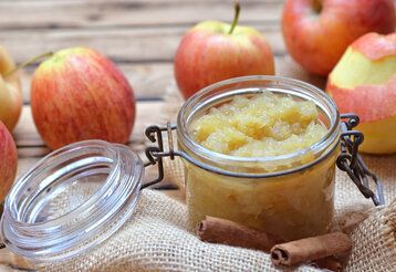 offenes Einmachglas mit Apfelmus auf Jutestoff und Holztisch, Äpfel drumherum und Zimtstangen