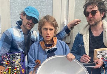 zwei Männer und eine Frau stehen mit Musikinstrumenten und Mischpulten nebeneinander