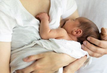 Baby wird von der Mutter im Arm gehalten und gestillt, die Hand des Vaters hält das Köpfchen