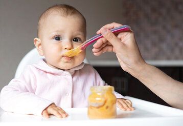 Baby wird mit Brei gefüttert und guckt dabei in die Kamera, der Arm einer erwachsenen Person führt den Löffeln zum Mund des Babys