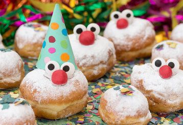Karnevalsgebäck Berliner mit Augen und Nasen und Hütchen, auf Tisch mit Konfetti und Luftschlangen
