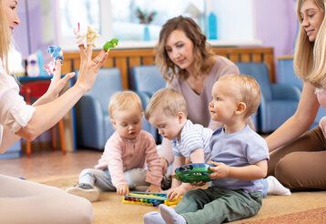 Drei Betreuerinnen und drei Kleinkinder sitzen auf dem Boden spielen zusammen, eine Betreuerin hat Fingerpuppen auf den Fingern