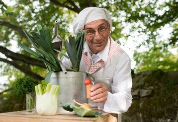 Der Märchenkoch in Kochuniform mit einem Kochtopf und Gemüse.