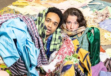 ein Mann und eine Frau liegen auf dem Boden, umgeben und überschüttet von Kleidungsstücken