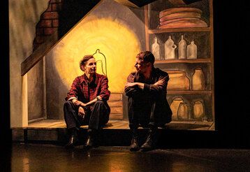 eine Schauspielerin und ein Schauspieler sitzen auf der Bühne, im Hintergrund eine gemalte Kulisse von einer Hütte