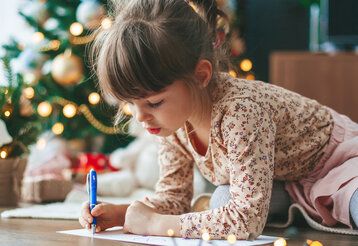 ein kleines Mädchen sitzt auf dem Boden und schreibt einen Brief, im Hintergrund: ein Weihnachtsbaum