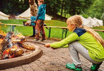 ein Mädchen hält einen Holzstock mit Brotteig ins Lagerfeuer und macht Stockbrot, im Hintergrund zwei Personen und Zelte auf einer Wiese