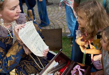 Eine Frau ist als Charlotte von Aschenberg verkleidet und zeigt zwei Mädchen ein Schriftstück im Kindermuseum Adlerturm