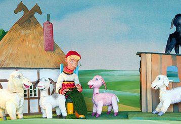 Eine Schäfer-Puppe sitzt strickend vor der Kulisse, umringt von Schafspuppen.