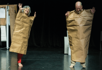 Zwei Schauspieler auf einer Bühne sind in große Papiertüten eingewickelt.