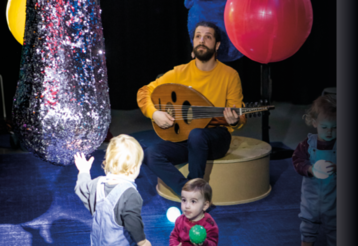 Ein Mann mit Ukulele spielt und singt inmitten von kleinen Kindern und Luftballons
