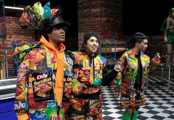 drei Schauspieler:innen auf einer Bühne, die wie ein Supermarkt aussieht, die Kleidung besteht aus Chipstüten