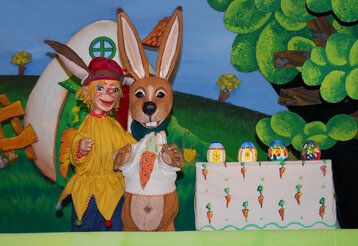 Kasper und Osterhase vor Puppenbühnen-Kulisse, mit bemalten Ostereiern