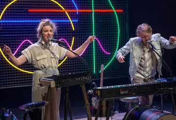 eine Musikerin und ein Musiker machen zusammen Musik, im Hintergund bunter Neonröhren