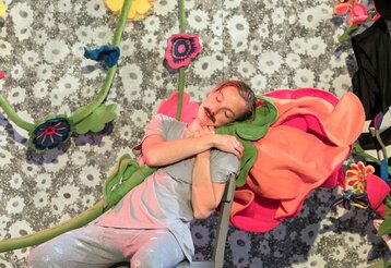 eine Schauspielerin mit einem Schnurrbart schläft auf einem Stuhl und hält dabei eine Stoffblume im Arm, im Hintergrund weitere Stoffblumen