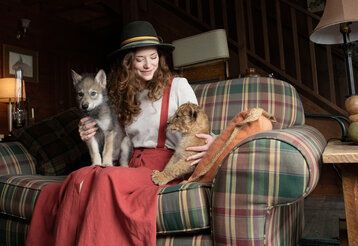 eine junge Frau mit Hut sitzt mit einem jungen Wolf und einem jungen Löwen auf einem karierten Sofa