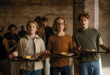 Drei Jungs stehen mit Tabletts, auf denen Teller mit Essen sind, in einem dunklen Gewölbe