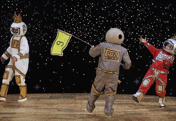Drei als Astronauten verkleidete Schauspieler auf einer Bühne, der in Astronaut in der Mitte hält eine Flagge, auf der eine 9 steht, in den Händen, im Hintergrund Weltall mit Sternen