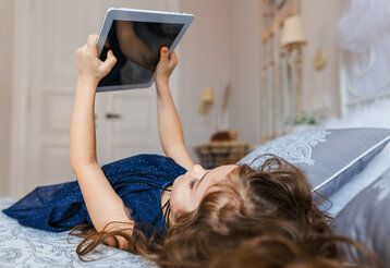 ein kleines Mädchen liegt auf dem Bett und hält ein Tablet in den Händen