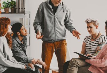 fünf Erwachsene sitzen zusammen im Gespräch, ein Mann in der Mitte steht und redet